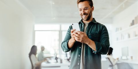 Business man reading a message on a mobile phone in an office. Male business professional standing in a co-working office.